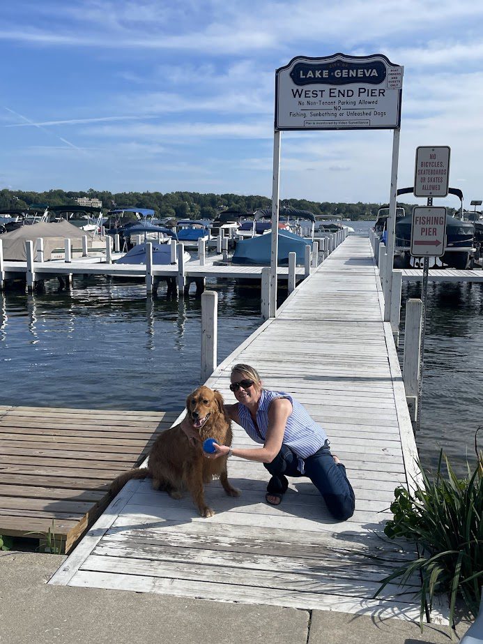 Two women sitting on a dock with a dog by the water under a sunny sky.