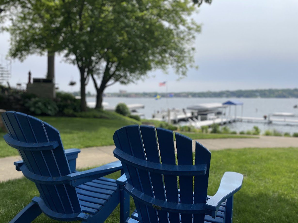 Blue Adirondack chairs overlooking a peaceful lakeside with boats and greenery.