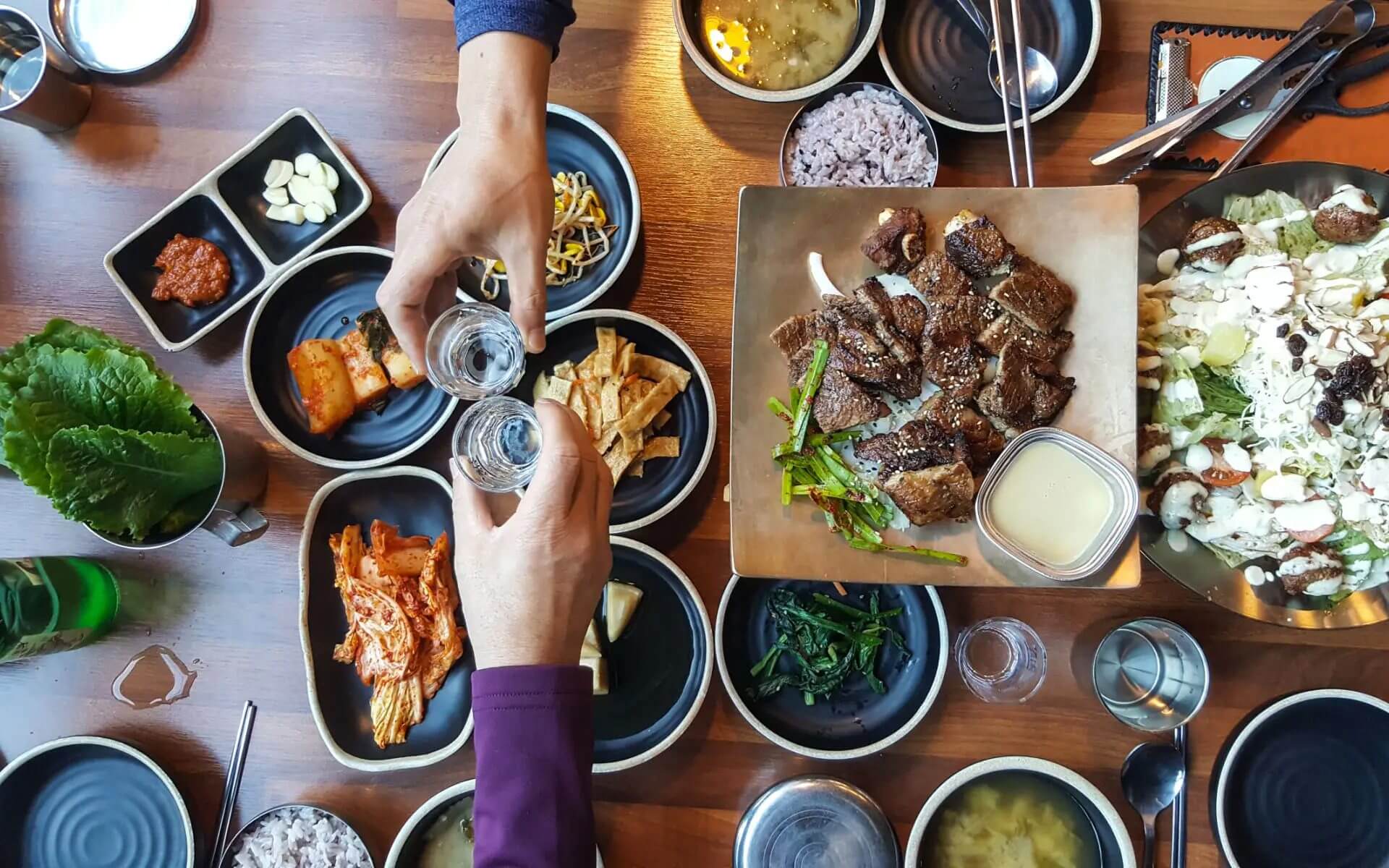 Two people clinking glasses over a Korean BBQ meal with various side dishes.