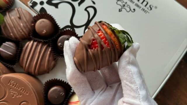 Gloved hand holding a chocolate-covered strawberry from Kilwin's Chocolates.