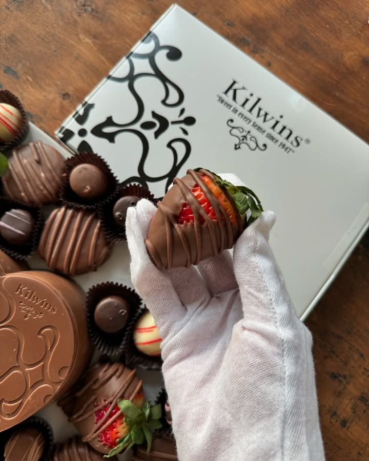 Gloved hand holding a chocolate-covered strawberry from Kilwin's Chocolates.