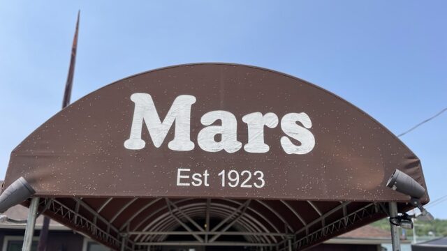 A brown awning with 'Mars Est 1923' written on it under a clear blue sky.