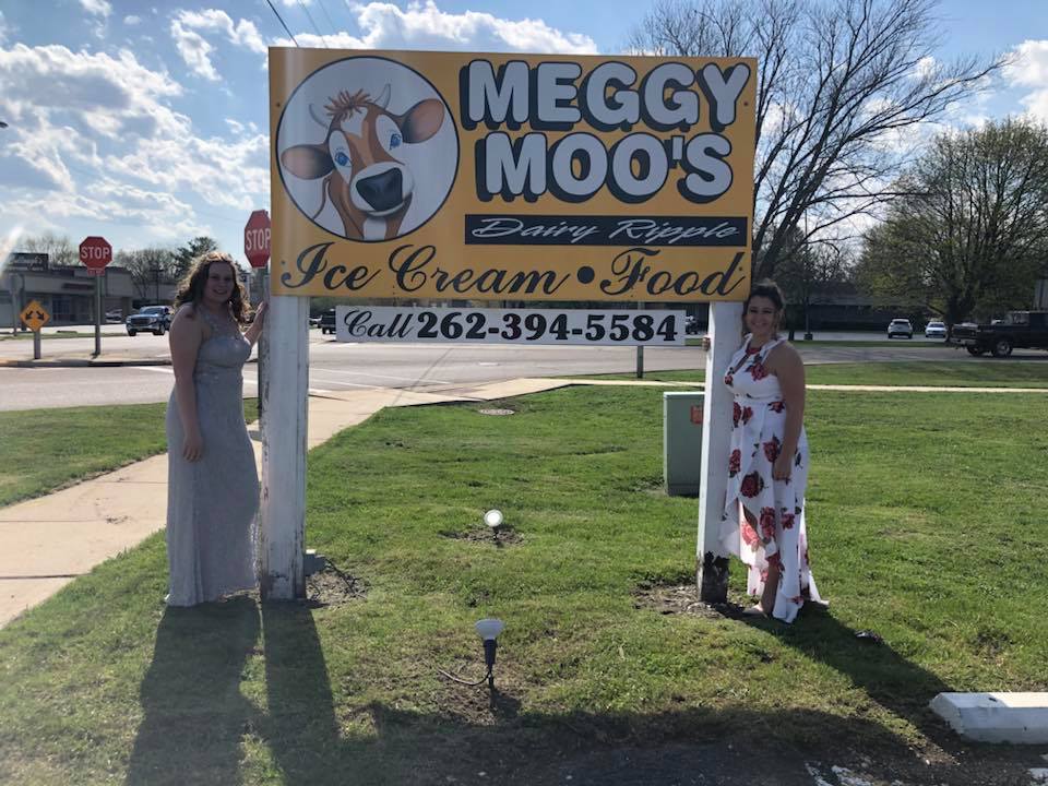 Two women posing beside a Meggy Moo's Ice Cream & Food sign outdoors.