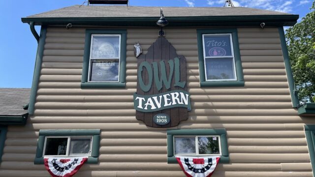 The Owl Tavern sign hangs on a tan building with patriotic bunting below.