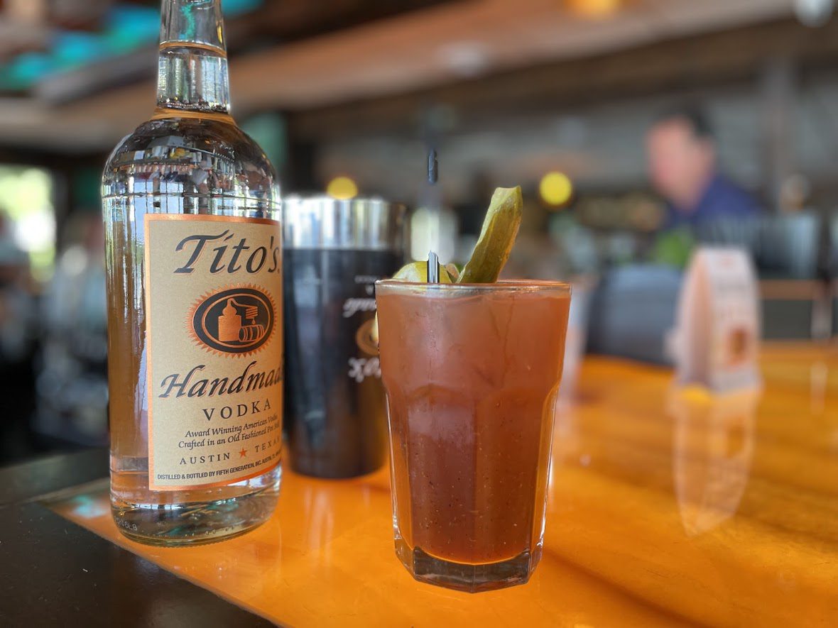 A Bloody Mary cocktail with a Tito's Handmade Vodka bottle on a wooden bar counter.