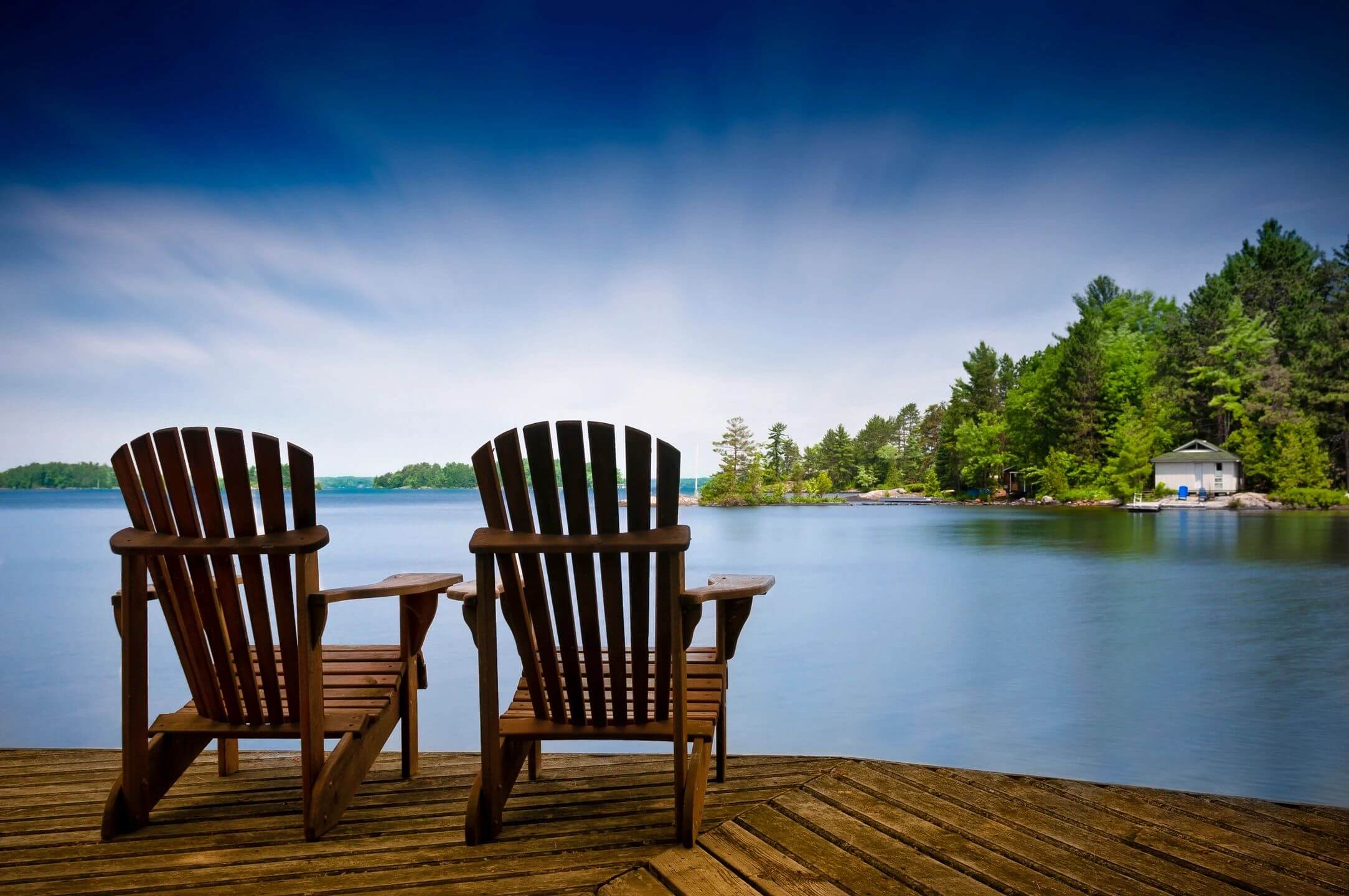 Two wooden chairs on a dock overlooking a calm lake and forest.