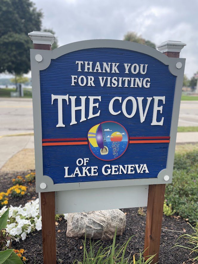 Welcome sign for The Cove of Lake Geneva with a decorative emblem.