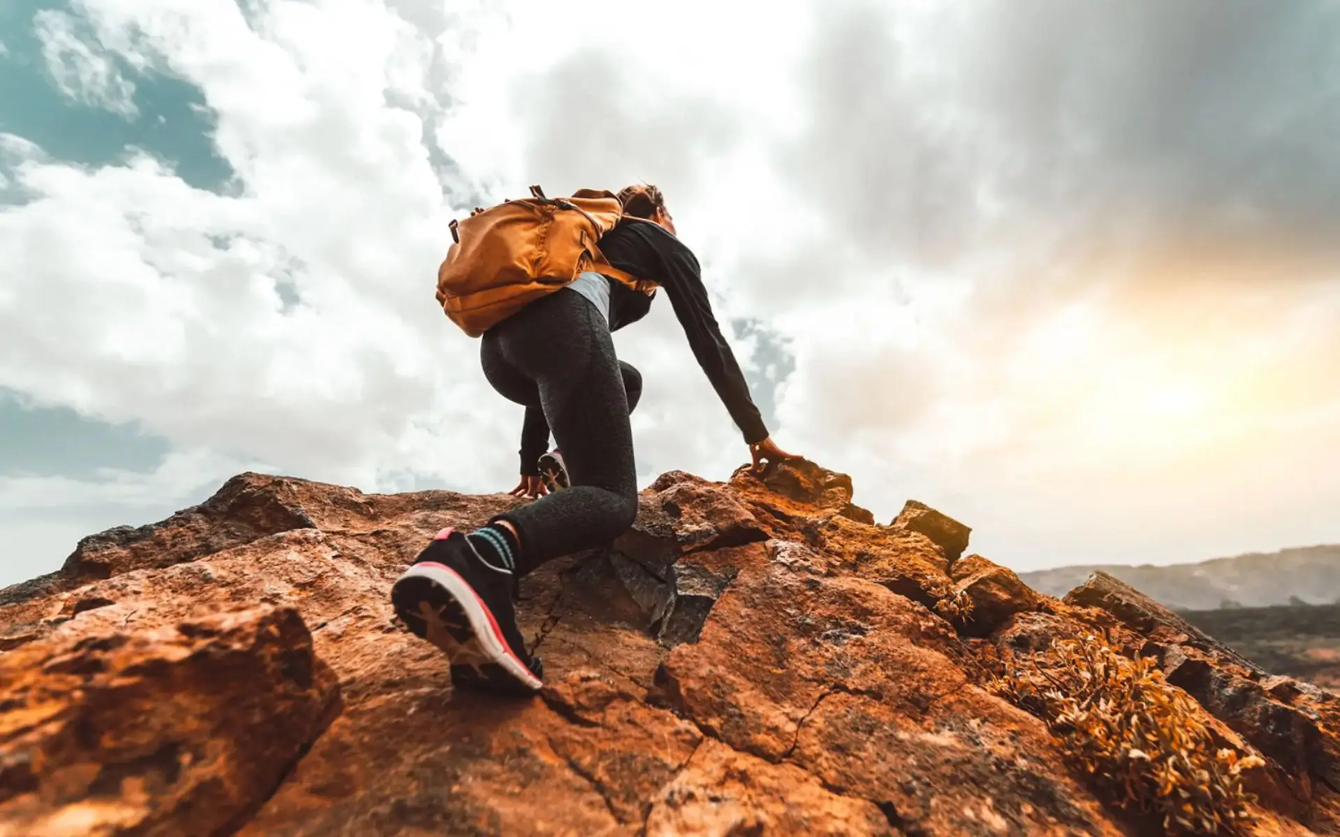 Person climbing rocky terrain with an orange backpack under a cloudy sky.